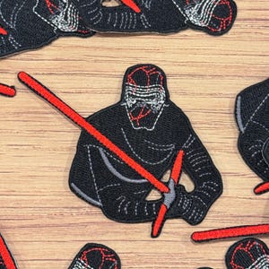 Puede incluir: Parche bordado en negro y rojo de un personaje de Star Wars, Kylo Ren, sosteniendo dos sables de luz rojos.