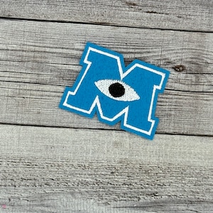 May include: Blue felt patch with a white outline in the shape of the letter "M". A white eye with a black pupil is centred on the patch.