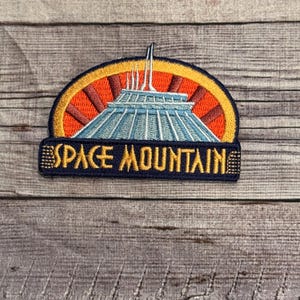 May include: Embroidered patch featuring the Space Mountain attraction. The patch has a blue and white building design with an orange and yellow background. The words "SPACE MOUNTAIN" are embroidered in gold.