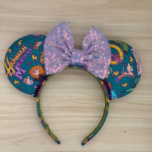 May include: A teal headband with two round ears featuring cartoon characters and the text "Hannah Montana". A large, sequined lavender bow sits between the ears. The headband is decorated with a green and purple pattern.