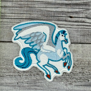 Puede incluir: Un parche bordado que representa un caballo alado con cola azul. El caballo está mirando hacia el lado derecho de la imagen y es de color azul y blanco.