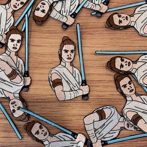 May include: Embroidered patches featuring a character from a science fiction film. The patches depict a person in a white and tan outfit, holding a blue lightsaber. The patches are arranged on a wooden surface.
