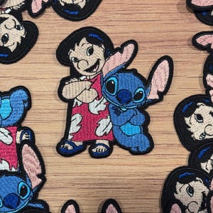 May include: Embroidered patches featuring Lilo and Stitch characters. The patches show Lilo in a red dress with white floral designs, hugging Stitch, a blue alien. The patches are on a wooden surface.