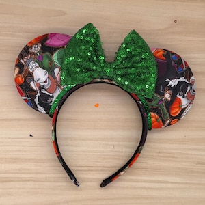 Puede incluir: Una diadema con temática de Halloween con orejas de ratón. Las orejas presentan una colorida impresión de personajes de dibujos animados y calabazas. Un gran lazo verde brillante con lentejuelas se encuentra en la parte superior de la diadema.