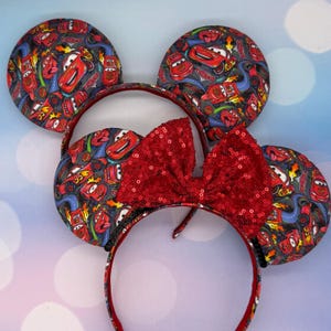 May include: Two sets of mouse ear headbands. The ears are round and feature a pattern of red, blue, and yellow cars on a gray background. A large red sequined bow sits between the ears. The headband is red.