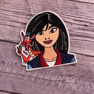 May include: Embroidered patch featuring Mulan from Disney's Mulan, with her black hair pulled back and a red dragon on her shoulder. The dragon has a red and white body with a blue and white horn.