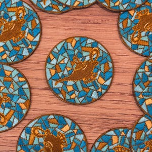 May include: Circular embroidered patches featuring a brown genie lamp design. The patches have a mosaic-like background in shades of blue and orange. The patches are arranged on a wooden surface.