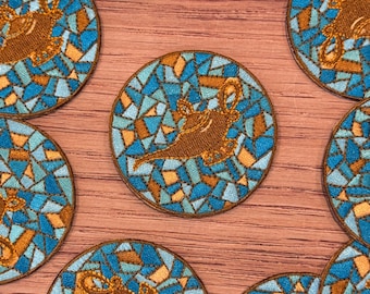 May include: Circular embroidered patches featuring a brown genie lamp design. The patches have a mosaic-like background in shades of blue and orange. The patches are arranged on a wooden surface.