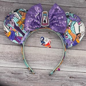 Puede incluir: Orejas de ratón de tela morada y verde azulado con un lazo de lentejuelas moradas. Las orejas presentan un estampado de personajes de la película de Disney "La Princesa y el Sapo". El texto "PUT THE  GECKO  BACK ON!" es visible en una pegatina.