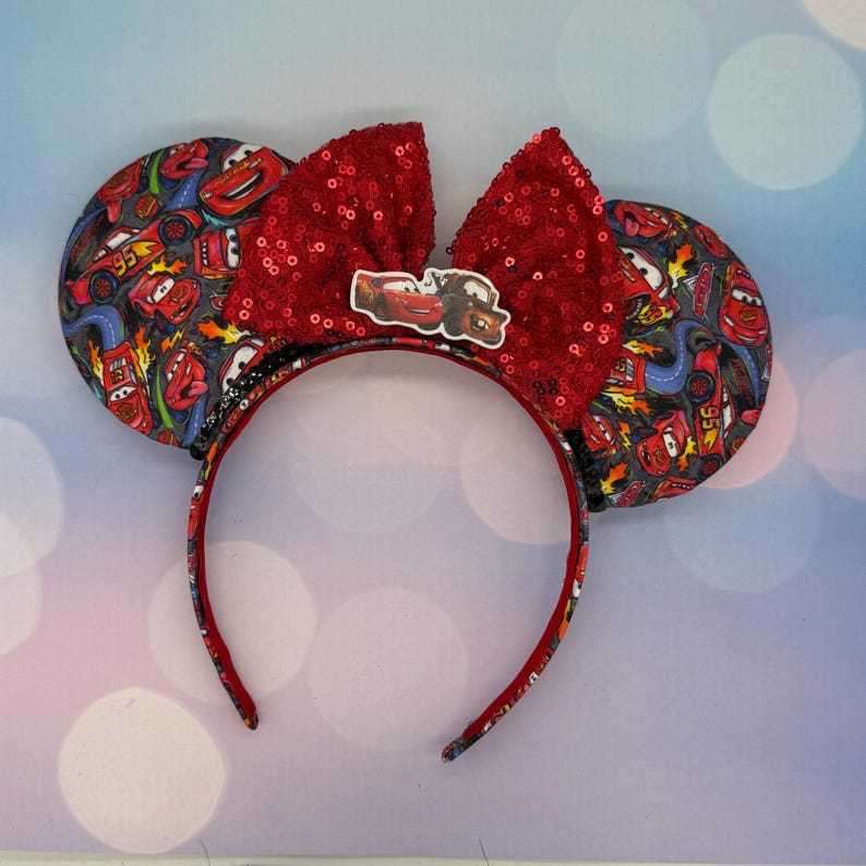 May include: A pair of themed mouse ears featuring a red sequined bow and ear details with a Cars movie print. The headband is red with a Cars character emblem in the center.