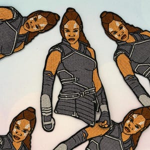 May include: Embroidered patches depicting a warrior woman with brown hair and skin, dressed in a grey and black outfit. The patches show detailed facial markings and armour-like clothing. These patches are likely for clothing or accessories.