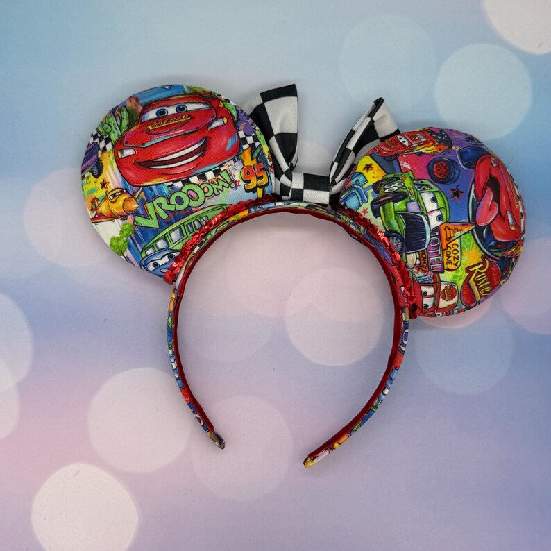 May include: A pair of mouse ears featuring a Cars theme. The ears are round and covered in a colorful fabric with images of Lightning McQueen and other characters. A black and white checkered bow sits between the ears, and the headband is red.
