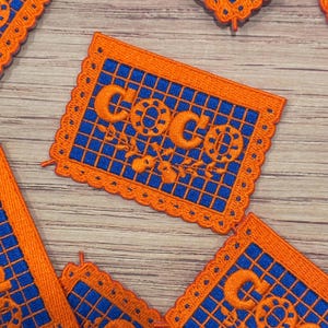 May include: Embroidered patches featuring the word "COCO" in orange, set against a blue grid. The patches have an orange scalloped border and floral details. Several patches are visible on a wooden surface.
