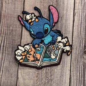 May include: Blue Stitch character from the Disney movie Lilo & Stitch reading a book with three small Stitch characters. The book is open and has colorful illustrations.