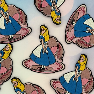 May include: Multiple embroidered patches featuring a character with blonde hair wearing a blue dress and white apron, standing inside a pink and brown teacup. The character is depicted in various orientations, showcasing detailed stitching and black outlines.