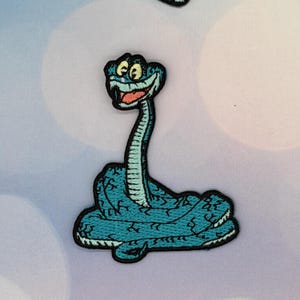 Pode incluir: Patch bordado com uma cobra de desenho animado em tons de azul-petróleo e azul. A cobra tem um rosto sorridente com boca vermelha e olhos amarelos. O patch tem uma borda preta e é para roupas ou acessórios.