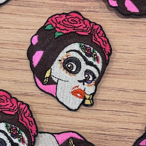 May include: Embroidered patch featuring a stylized skull with a floral crown and pink accents. The skull has detailed facial features, including large eyes, red lips, and gold earrings. The patch is designed with a Day of the Dead theme.