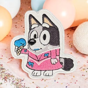 Puede incluir: Parche bordado para planchar con un perro de dibujos animados que lleva un vestido rosa y sostiene un cono de helado azul. El perro tiene pelo blanco y negro y una lengua azul.