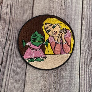 May include: Embroidered patch featuring a cartoon of Rapunzel from Disney's Tangled with her chameleon friend Pascal. Rapunzel has long blonde hair and is wearing a pink dress. Pascal is green and wearing a pink dress. The patch is circular and has a brown background.