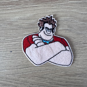 Op de afbeelding: Geborduurde patch met een cartoonfiguur met bruin haar, een rood shirt en een witte achtergrond. Het personage glimlacht en heeft zijn armen gekruist.