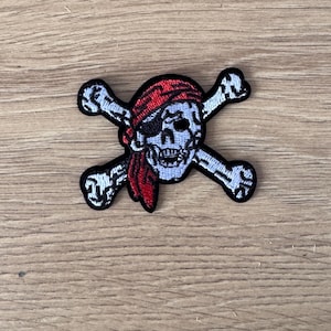 Może przedstawiać: Biała czaszka z czerwoną bandaną i opaską na oko, skrzyżowana z dwiema białymi kośćmi. Czaszka i kości są wyszyte czarną nicią.