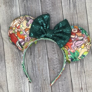 May include: A pair of mouse ears with a green sequin bow. The ears feature a colorful Jungle Book print. The headband is decorated with a floral pattern and green leaves.