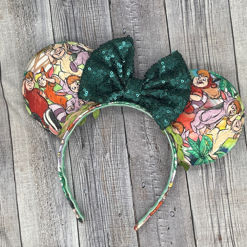 May include: A pair of mouse ears with a green sequin bow. The ears feature a colorful Jungle Book print. The headband is decorated with a floral pattern and green leaves.