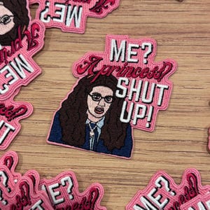 Puede incluir: Parche bordado rosa con el texto "¿YO? ¿Una princesa? ¡CÁLLATE!" y una imagen de dibujos animados de una mujer con gafas. El parche tiene un borde rosa y está rodeado de otros parches.