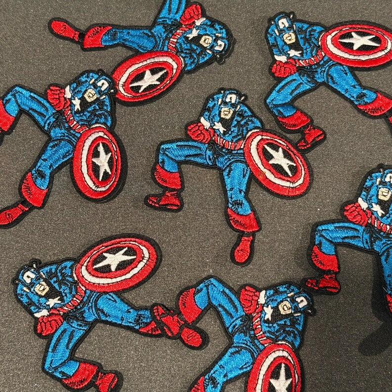 May include: Embroidered Captain America patches in various action poses. The patches depict the character in a blue and red costume, holding his shield with a white star on a red background. The patches are set against a grey background.