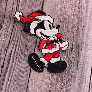 Op de afbeelding: Een geborduurde strijk-patch van een cartoonmuis die verkleed is als kerstman. De muis draagt een rood pak met witte versieringen, een witte hoed met een groene hulsttak en zwarte schoenen.
