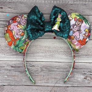 May include: A Peter Pan-themed headband with large, sequined green bow and Peter Pan character charm. The ears feature a colorful print with characters from the movie. The headband is decorated with a floral pattern.