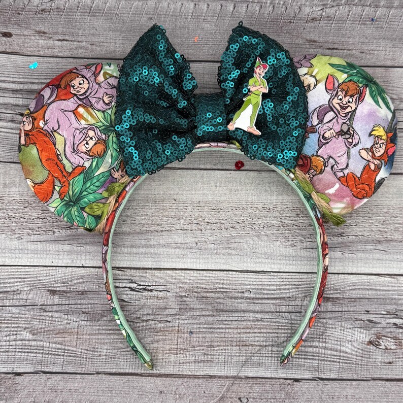 May include: A Peter Pan-themed headband with large, sequined green bow and Peter Pan character charm. The ears feature a colorful print with characters from the movie. The headband is decorated with a floral pattern.