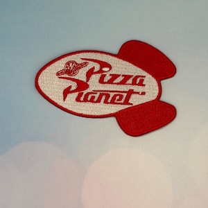 May include: Embroidered patch featuring the words "Pizza Planet" in red, stylized lettering. The patch is shaped like a red rocket with a white oval in the center. A logo of a planet and a spaceship is above the word "Pizza".