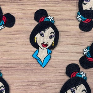 May include: A set of embroidered iron-on patches featuring Mulan from the Disney movie Mulan. The patches are colorful and feature Mulan's face, hair, and outfit.