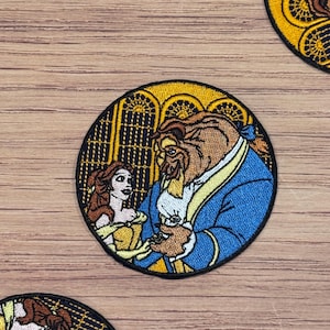 May include: A round embroidered patch featuring Belle and the Beast from Disney's Beauty and the Beast. The patch is yellow, blue, and brown with a black outline. Belle is wearing a yellow dress and the Beast is wearing a blue suit.