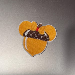 Puede incluir: Una bellota marrón y amarilla con una tapa marrón y un tallo rojo y blanco. La bellota está bordada en una tela blanca.