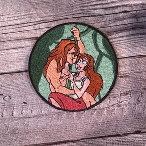May include: Embroidered patch featuring a cartoon couple, a man with brown hair and a woman with long red hair, holding onto a vine in a jungle setting.