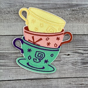 May include: A stack of three teacups with different patterns. The top cup is yellow with white stars, the middle cup is pink with white snowflakes, and the bottom cup is green with white stars and a swirl design. The cups are on a wooden background.