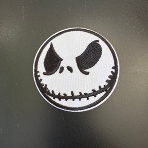 Könnte beinhalten: Gestickter Aufnäher mit dem Gesicht von Jack Skellington aus The Nightmare Before Christmas. Das Design hat einen weißen Hintergrund mit schwarzen gestickten Details für Augen, Mund und Augenbrauen. Rund.