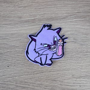 May include: Purple embroidered patch of a cartoon cat with a mischievous expression, holding a pink bottle. The cat has black eyes and a white mouth with sharp teeth.