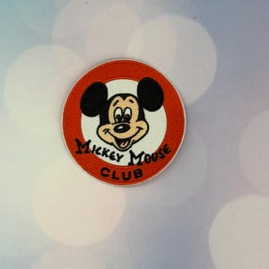 May include: A circular embroidered patch featuring the face of Mickey Mouse centered on a white background. The patch has a wide orange border with the black text Mickey Mouse Club printed in a curved font along the bottom half of the design.