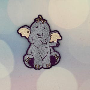 May include: Embroidered patch of a cartoon elephant in grey, with light beige ears and accents. The elephant is sitting, with purple details on its feet and edges. The patch has a smiling expression and is suitable for clothing or accessories.