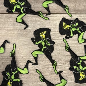 May include: Embroidered patches featuring a cartoon character in a dynamic pose. The patches are primarily black with green accents, depicting a female character with a determined expression. The patches are likely for clothing or accessories.