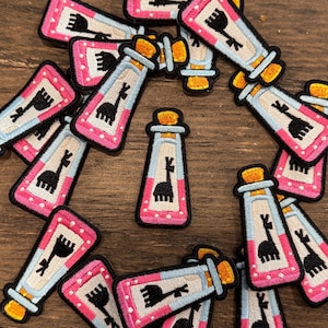 May include: A collection of embroidered patches featuring a potion bottle design with a llama silhouette. The patches have a pink border with white polka dots, a light blue center, and a cork top. The llama is black.
