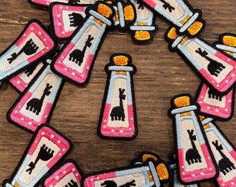 May include: A collection of embroidered patches featuring a potion bottle design with a llama silhouette. The patches have a pink border with white polka dots, a light blue center, and a cork top. The llama is black.