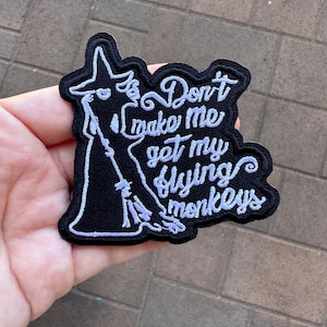 Puede incluir: Un parche bordado negro con una silueta blanca de una bruja sosteniendo una escoba. El texto "Don't make me get my flying monkeys" está bordado en blanco.