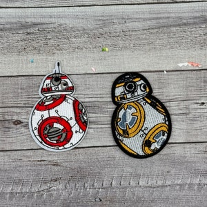 May include: Two embroidered patches of the Star Wars character BB-8. One patch is white with red and gray details, the other is white with yellow and gray details.