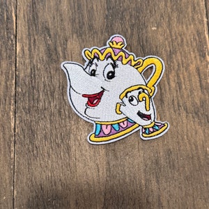 Pode incluir: Etiqueta bordada para passar a ferro com a Sra. Bule e Chip da Bela e o Monstro da Disney. A Sra. Bule é um bule branco com uma tampa rosa e amarela e uma borda roxa e amarela. Chip é uma pequena chávena com uma pega amarela e uma borda azul.