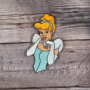 May include: Embroidered patch featuring Cinderella in her iconic blue gown and gloves, with blonde hair and a blue headband. The patch is on a wooden background.