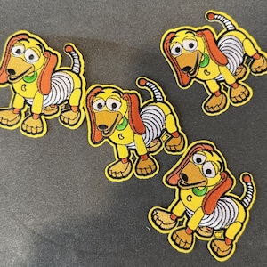 May include: Five embroidered patches featuring a cartoon dog with yellow, orange, and white details. The dog has floppy ears, a long body, and a curled tail. The patches are on a gray background.